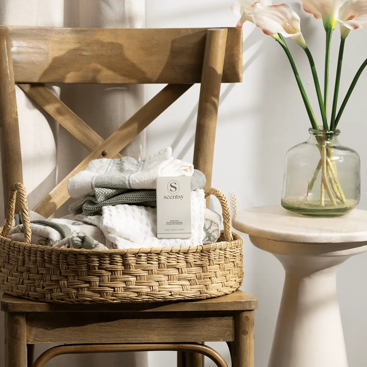 Woven wicker basket holding folded linens and a white Scentsy Clothesline Dryer Disks box on a wooden chair with cross-back design