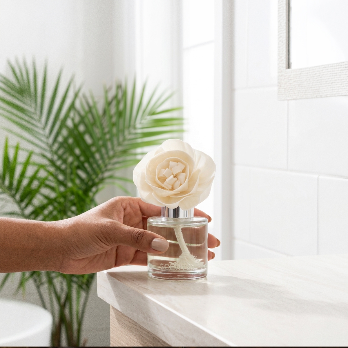 Cream rose-shaped Fragrance Flower with a glass diffuser base, the Amazon Rain - Buttercup Belle Fragrance Flower, held in a hand near a sunny window with potted palm foliage behind