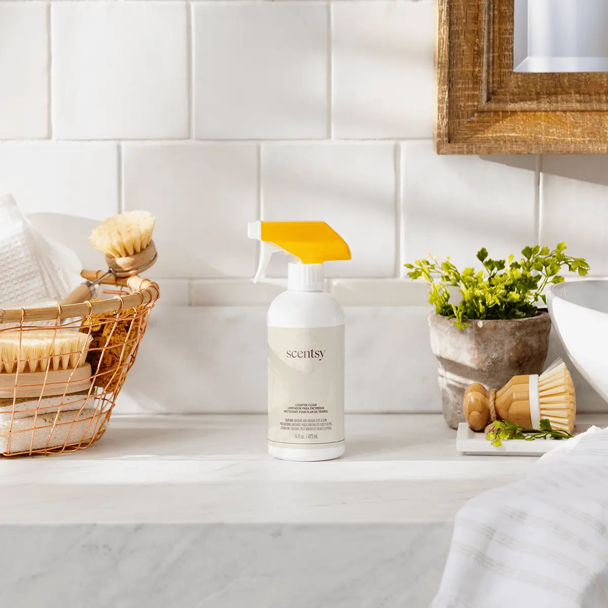 White spray bottle with yellow trigger and Scentsy branding, Squeeze the Day Counter Clean, displayed on a marble countertop beside cleaning brushes and potted herbs