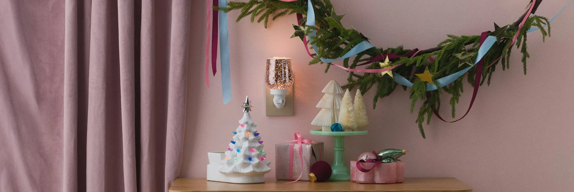 Pink Christmas scene with a white Scentsy tree warmer lit with coloured lights, a sparkling plug-in mini warmer, wrapped gifts and bottle-brush trees beneath a ribbon-tied garland