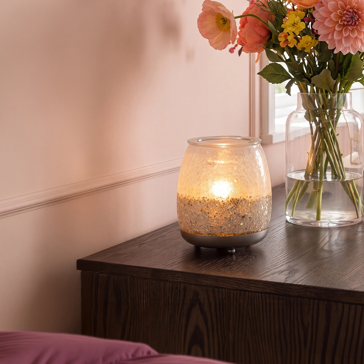 Lit Scentsy Warmer with a frosted glass upper half and glittery gold-bronze lower section, sitting on a dark wood dresser next to a vase of fresh flowers