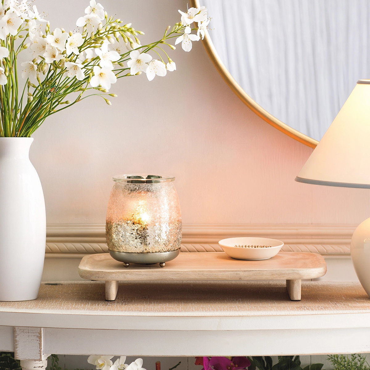 Textured glass warmer with gold speckled finish and metal base, lit from within, displayed on a wooden tray alongside white flowers and decor