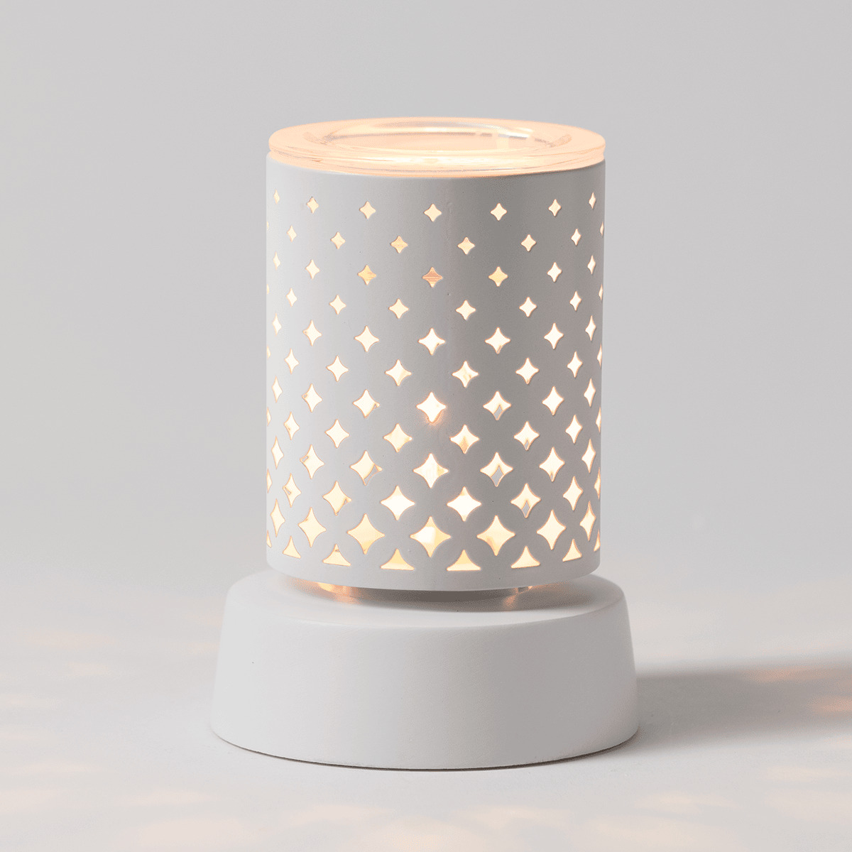White cylindrical ceramic warmer with a repeating four-pointed star cutout pattern that glows warmly when lit, sitting on a matching white cylindrical tabletop base, the Light From Within Mini Scentsy Warmer with Tabletop Base
