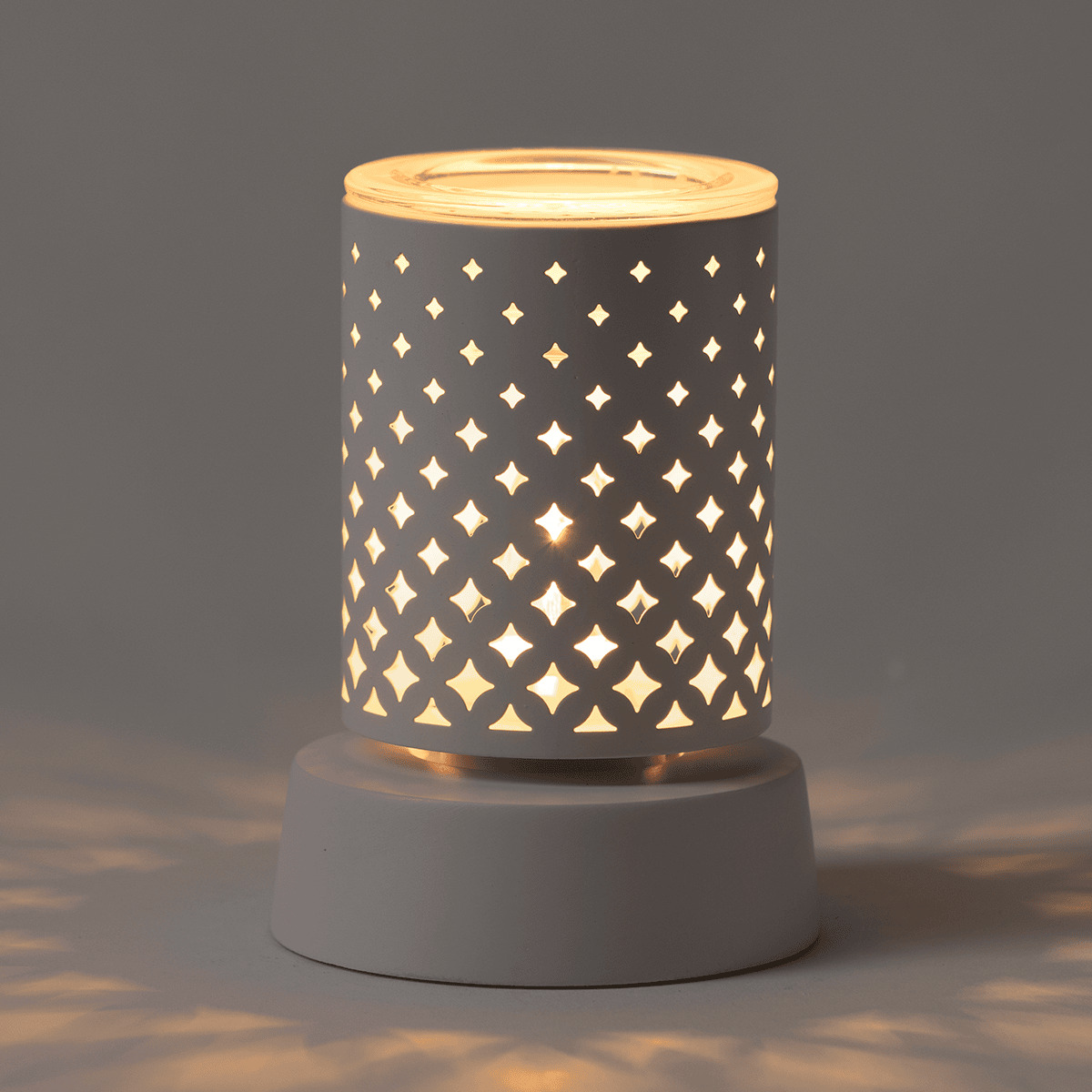 Cylindrical ceramic mini warmer with cut-out diamond pattern casting warm light, seated on a white ceramic base, the Light From Within Mini Scentsy Warmer with Tabletop Base
