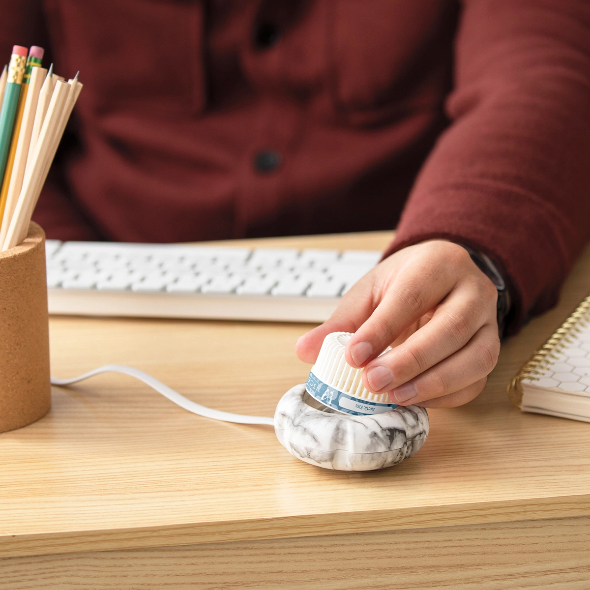 Hand holding a small marble-white diffuser with a ribbed top and blue band, sitting on a wooden desk next to pencils and notebooks