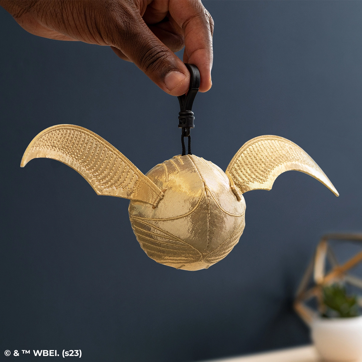 Hand holding a golden mesh plush toy with large bat-like wings and a spherical body, suspended by a black clip attachment