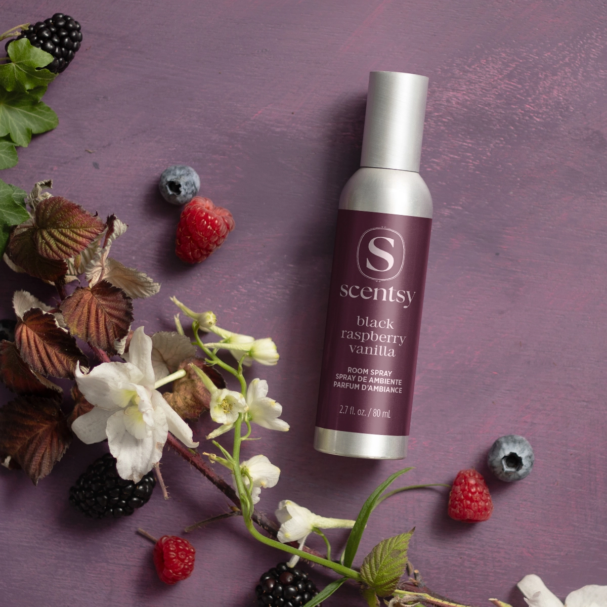 Spray bottle with silver cap and burgundy label showing the Scentsy logo and "black raspberry vanilla" text, surrounded by fresh raspberries, blackberries, blueberries and white flowers on a purple surface