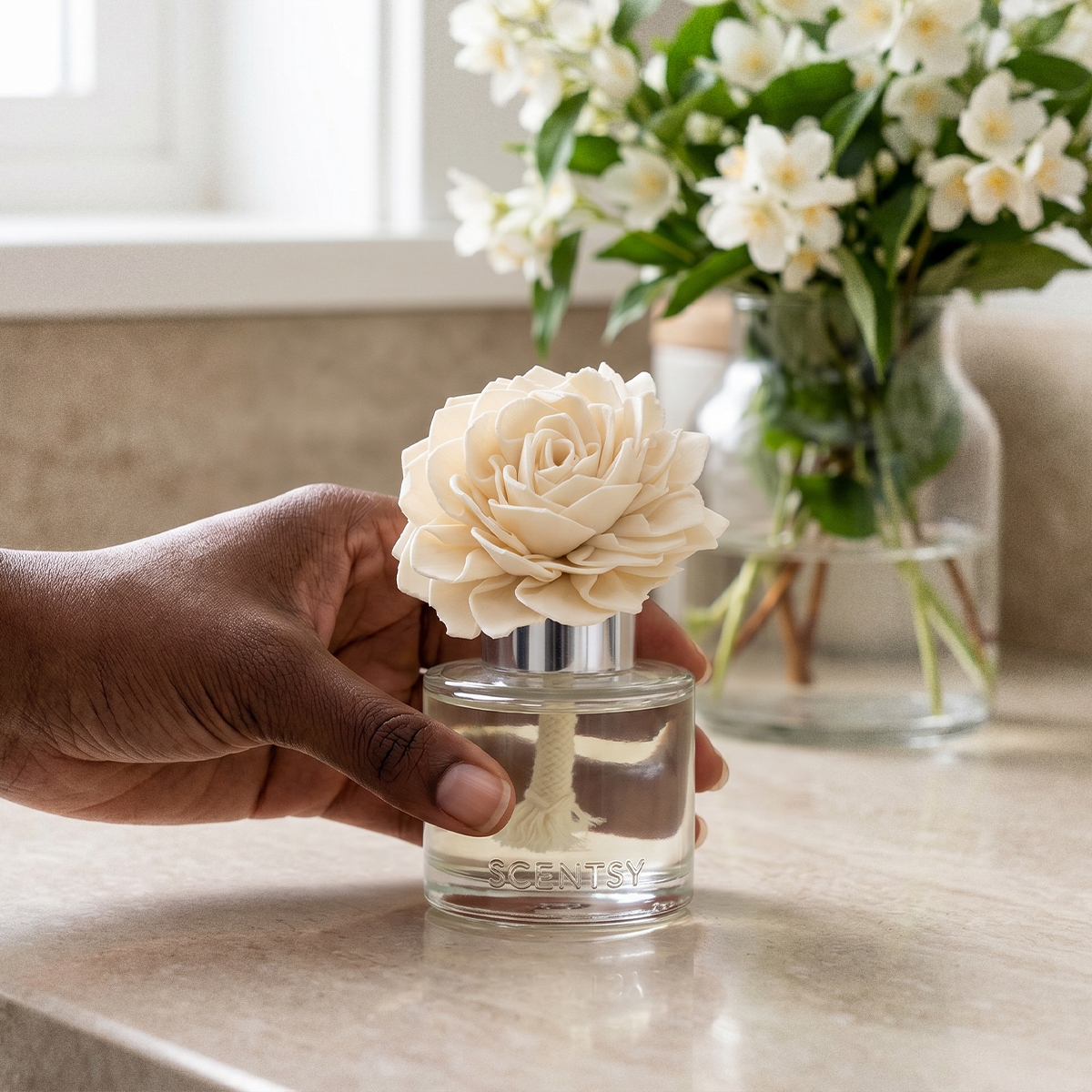 Cream dahlia-shaped Fragrance Flower with a cream resin stem inside a clear glass bottle marked "SCENTSY", held in hand above a kitchen counter with white flowers in a vase blurred behind