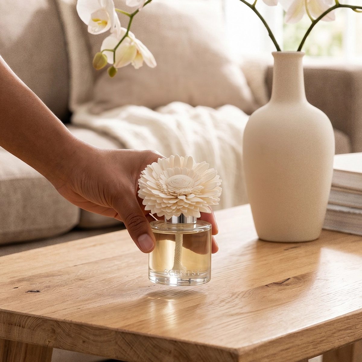 Hand holding a cream-coloured dainty daisy fragrance flower above a clear glass diffuser base filled with pale liquid, displayed on a wooden table with a cream ceramic vase of white orchids and neutral home décor in the background