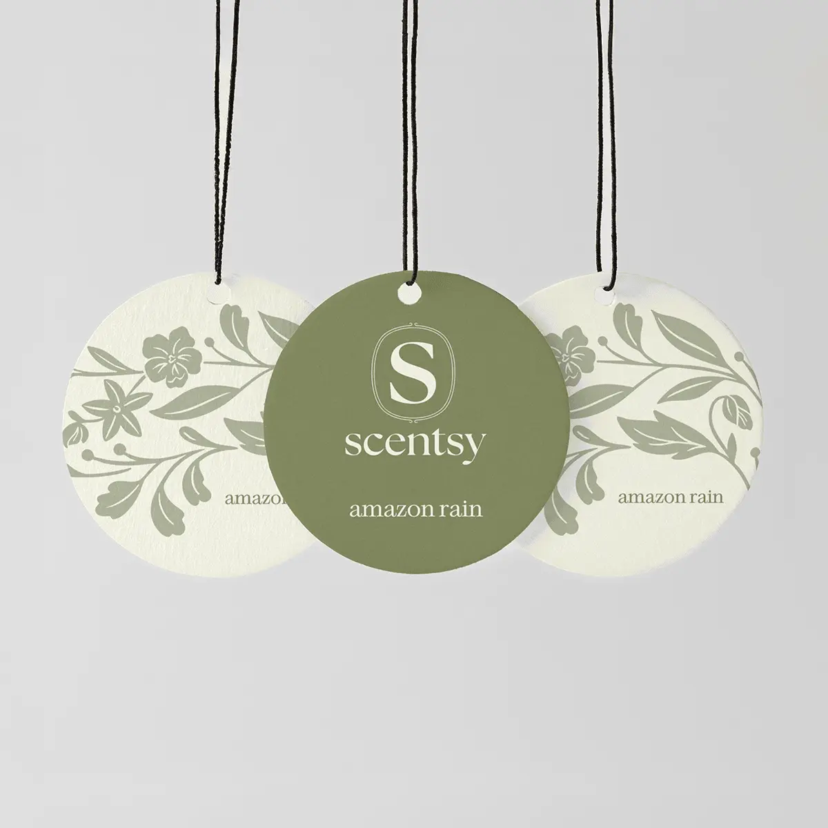 Three circular Scentsy Scent Circles with black strings: a sage-green centre circle and two cream circles with botanical leaf and flower designs, the Amazon Rain Scent Circle 3-pack