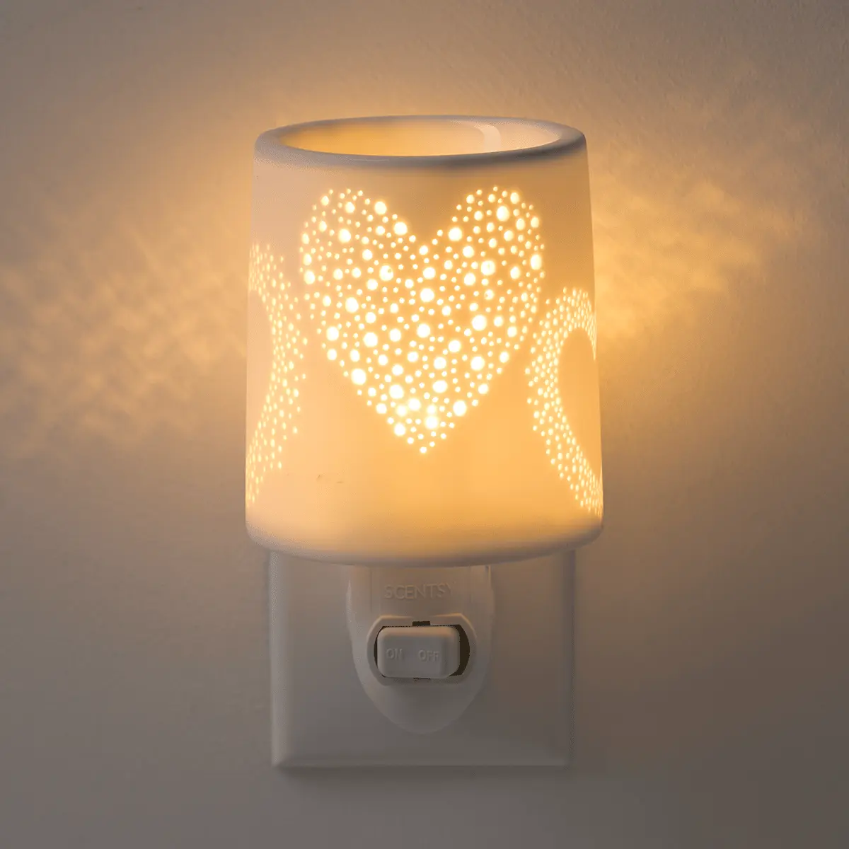 Cylindrical porcelain warmer glowing warm white, with a pierced heart design casting light patterns, mounted on a wall plug base