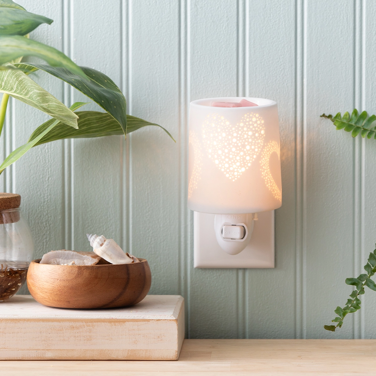 White cylindrical wall plug warmer with perforated heart cutout design casting warm light, the Heart to Heart Mini Scentsy Warmer