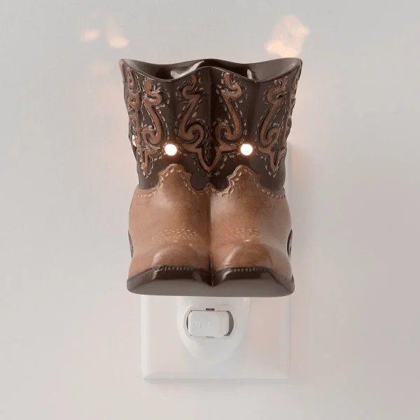 Two-tone ceramic cowboy boot warmer in tan and dark brown with ornate scrollwork detailing, lit from within, mounted on a white wall plug base, the Rodeo Mini Scentsy Warmer