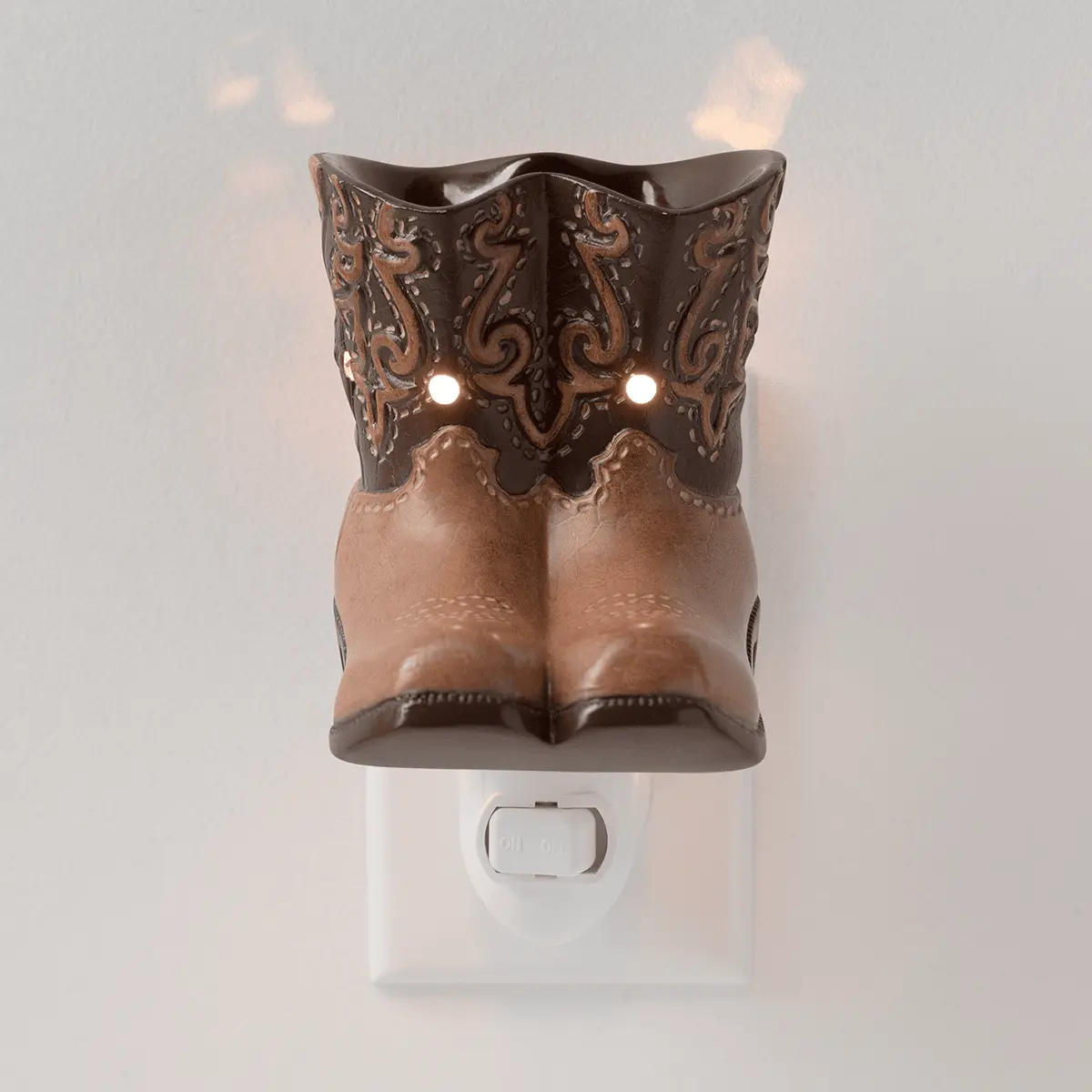 Two-tone ceramic cowboy boot warmer in tan and dark brown with ornate scrollwork detailing, lit from within, mounted on a white wall plug base, the Rodeo Mini Scentsy Warmer