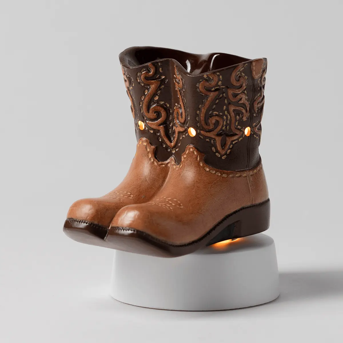 Brown and tan cowboy boot-shaped mini warmer with dark embossed scrollwork, sitting on a white ceramic base with warm light glowing from within