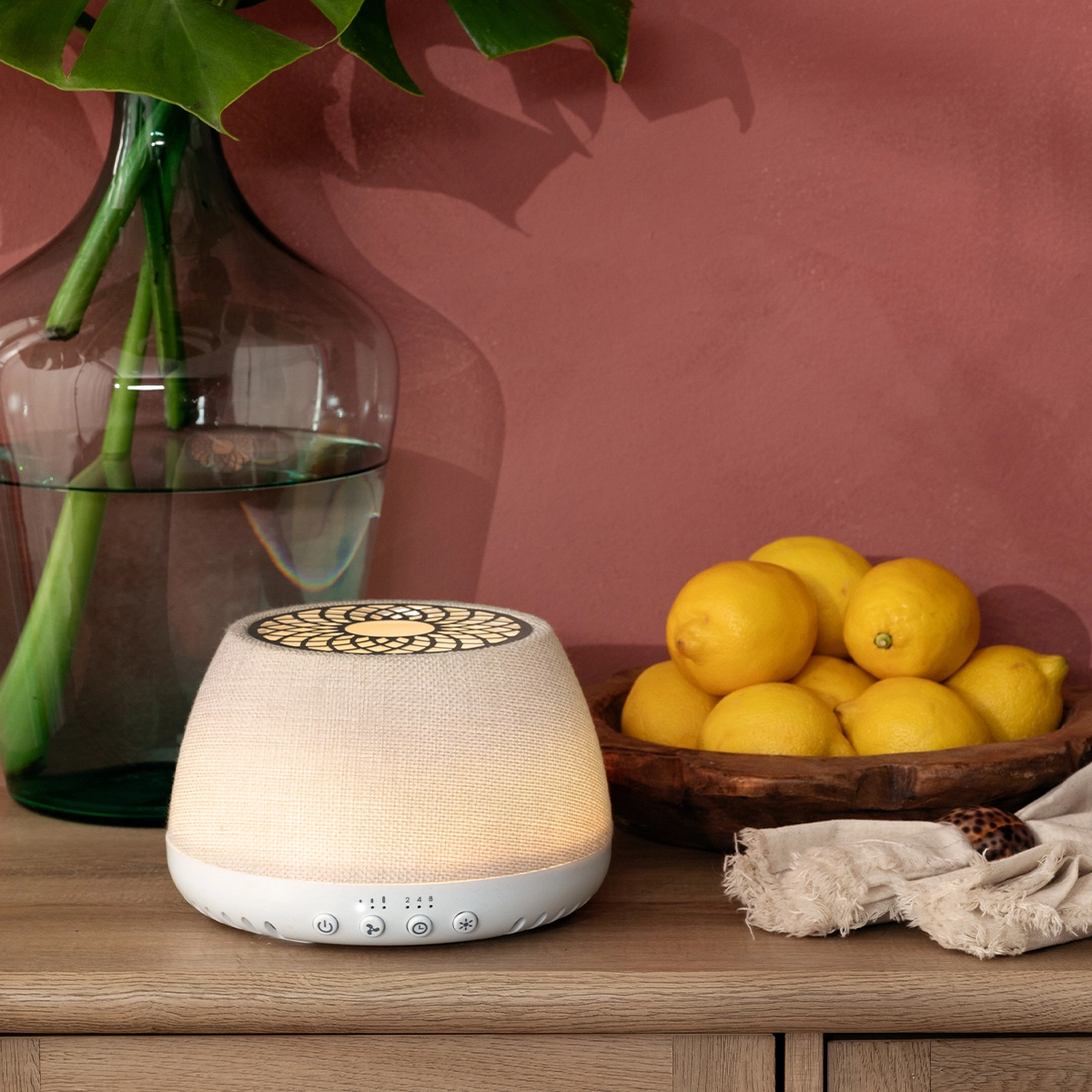 Cream linen-textured Scentsy Air Quad diffuser with white base and decorative circular pattern on top, displayed on a wooden shelf with lemons and fresh foliage