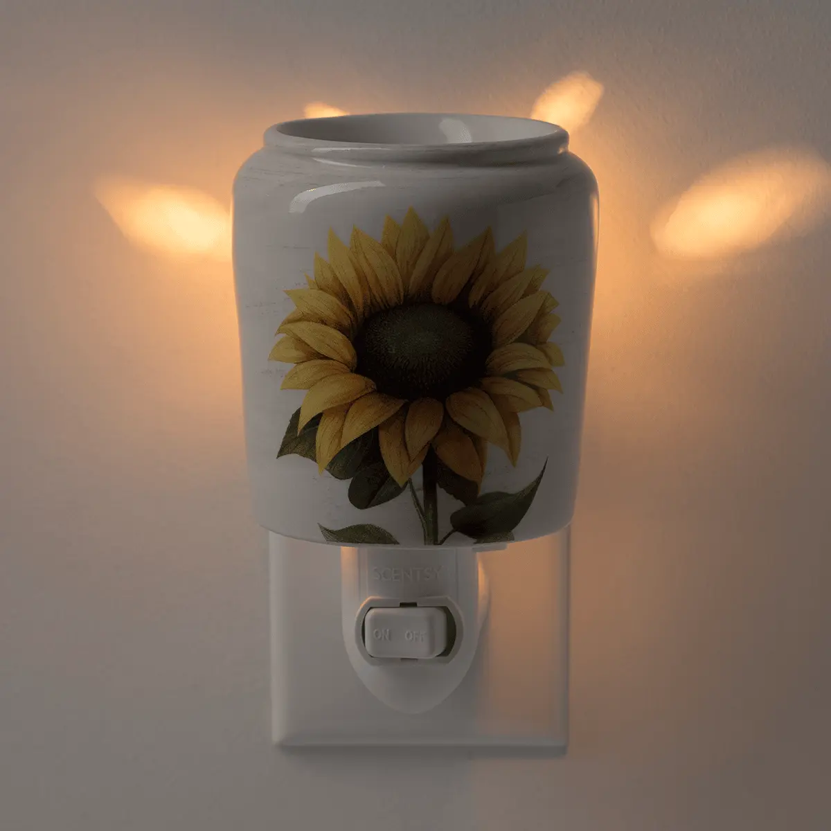 White ceramic mini warmer with a golden sunflower illustration and dark green leaves, mounted on a wall plug base with an on/off switch, glowing warmly when lit
