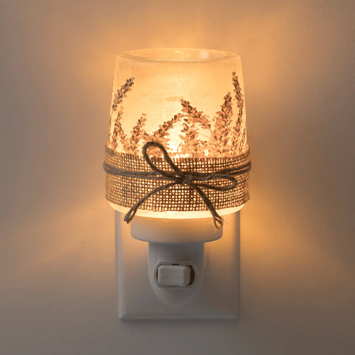 Lit cylindrical warmer with pressed lavender stems design, gold mesh band with black bow, mounted on white wall plug base