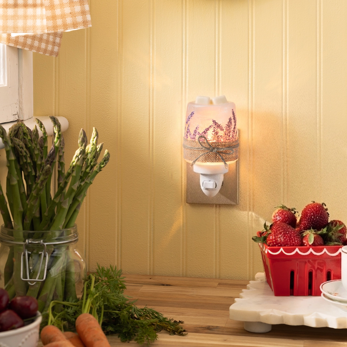 Pale pink plug-in warmer with lavender sprigs and burlap bow, glowing warmly on a kitchen wall surrounded by fresh vegetables and berries