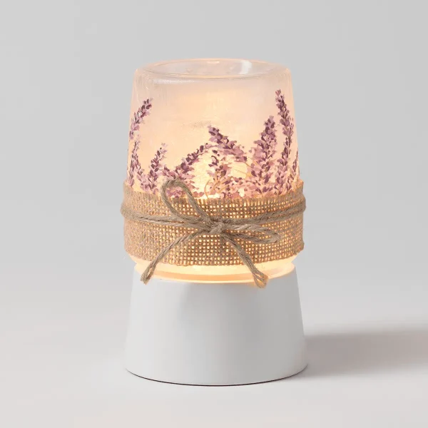 Frosted glass warmer with purple lavender sprigs printed around upper chamber, cream fabric bow and textured gold ribbon band, white ceramic base, the Lavender Fields Mini Scentsy Warmer with Tabletop Base, lit from within