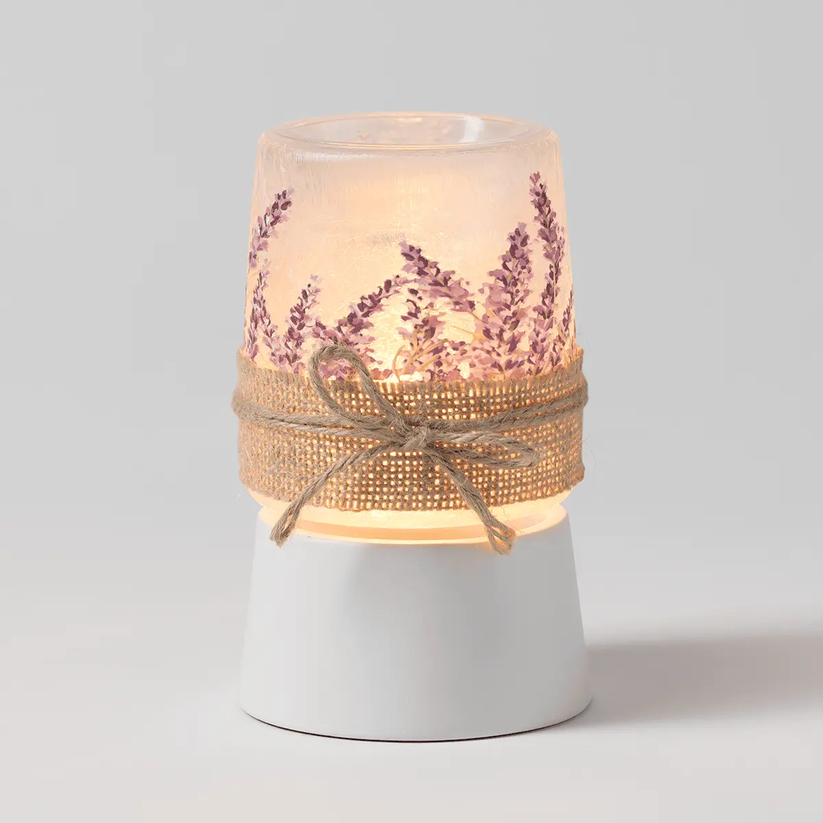 Frosted glass warmer with purple lavender sprigs printed around upper chamber, cream fabric bow and textured gold ribbon band, white ceramic base, the Lavender Fields Mini Scentsy Warmer with Tabletop Base, lit from within