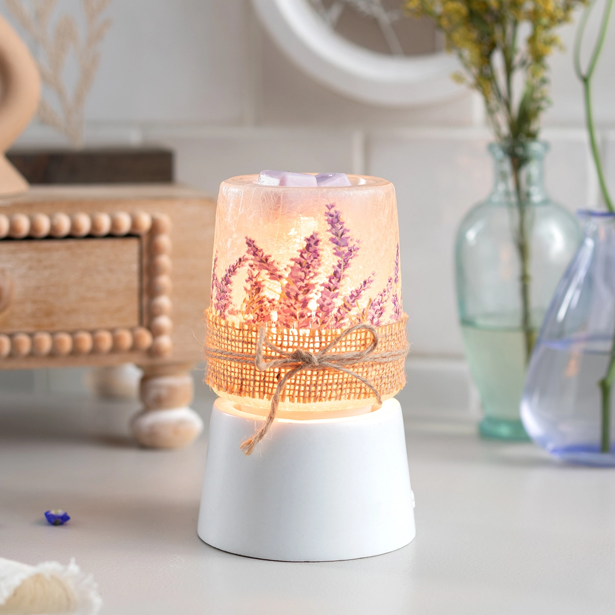 Cylindrical warmer with white ceramic base and printed shade depicting purple lavender stems on a cream background, wrapped with burlap ribbon and twine bow, glowing warmly when lit
