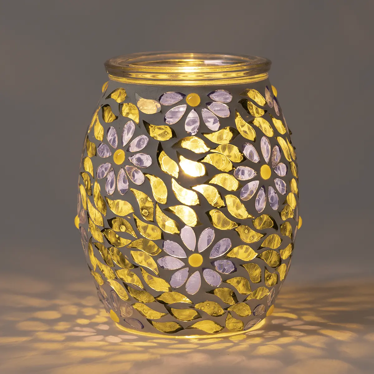 Barrel-shaped warmer with a grey metal cutout pattern of yellow leaves and purple flowers, illuminated from within, casting decorative shadows
