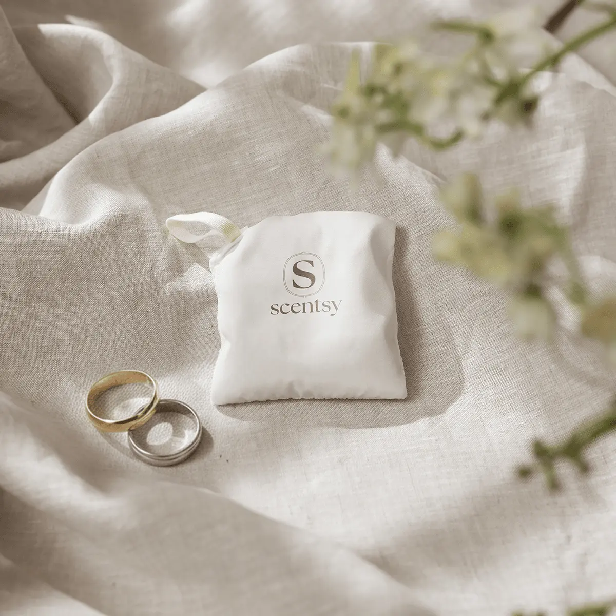 Small white fabric Scentsy Scent Pak with "S scentsy" embossed on it, nestled on cream linen beside two gold rings and white flowers