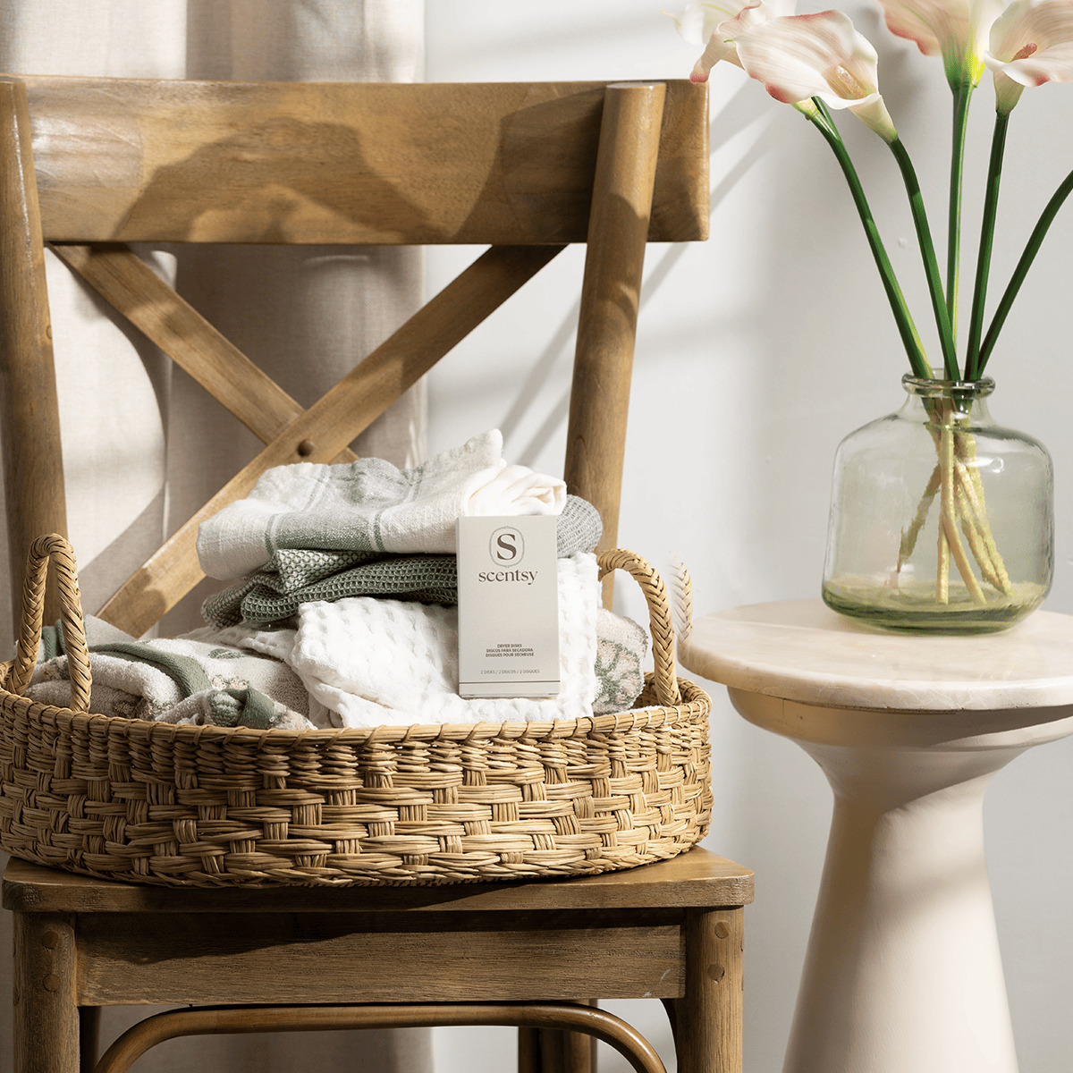 Woven basket holding folded textiles and a white box of Water Lily & Bergamot Dryer Disks, displayed on a wooden chair with rope handles beside a glass vase of pale pink flowers