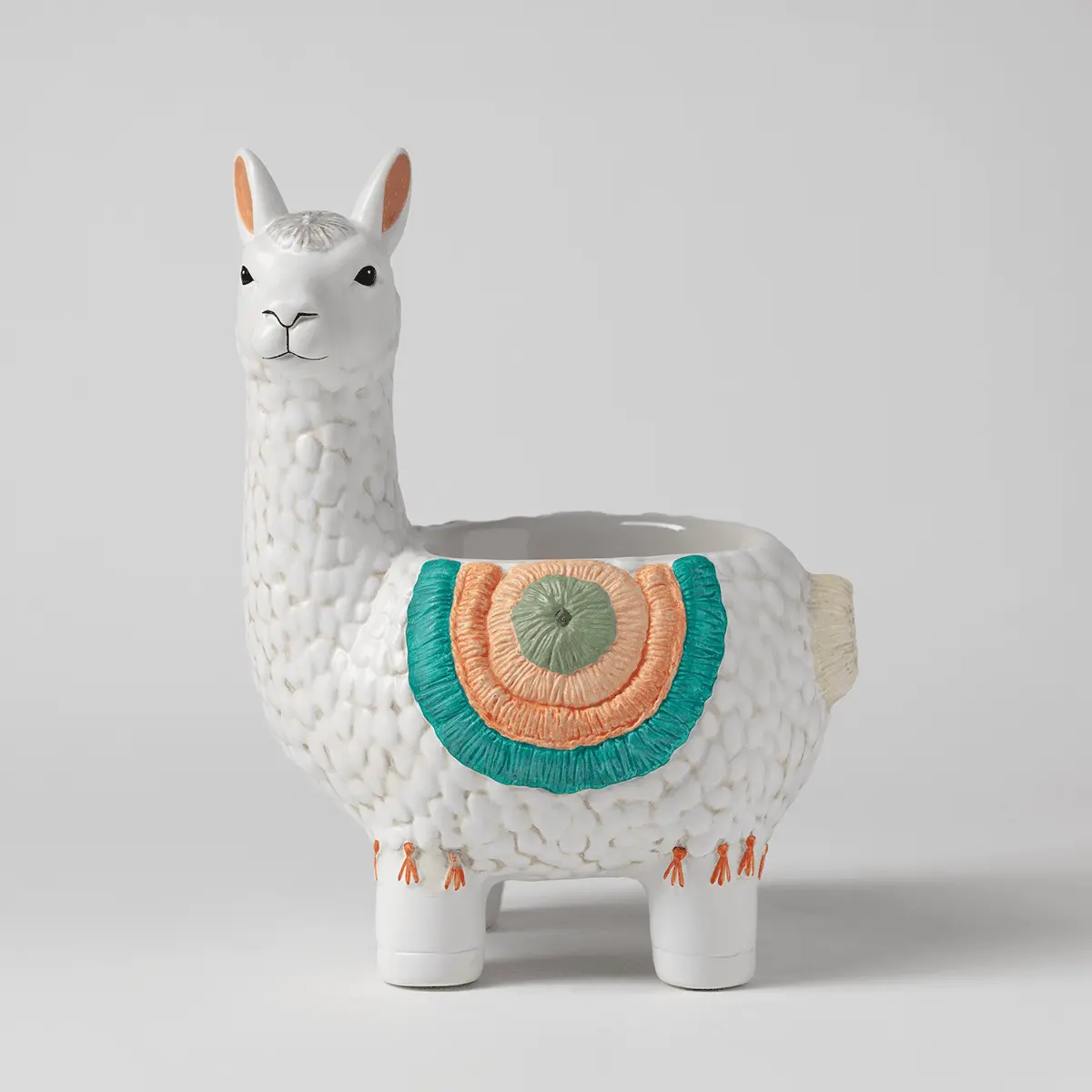 White ceramic llama-shaped warmer with textured wool detail, peach inner ears, black eyes and nose, a colourful geometric blanket in teal, orange and sage green across its back, and small orange figure details on its legs, the Llama Scentsy Warmer