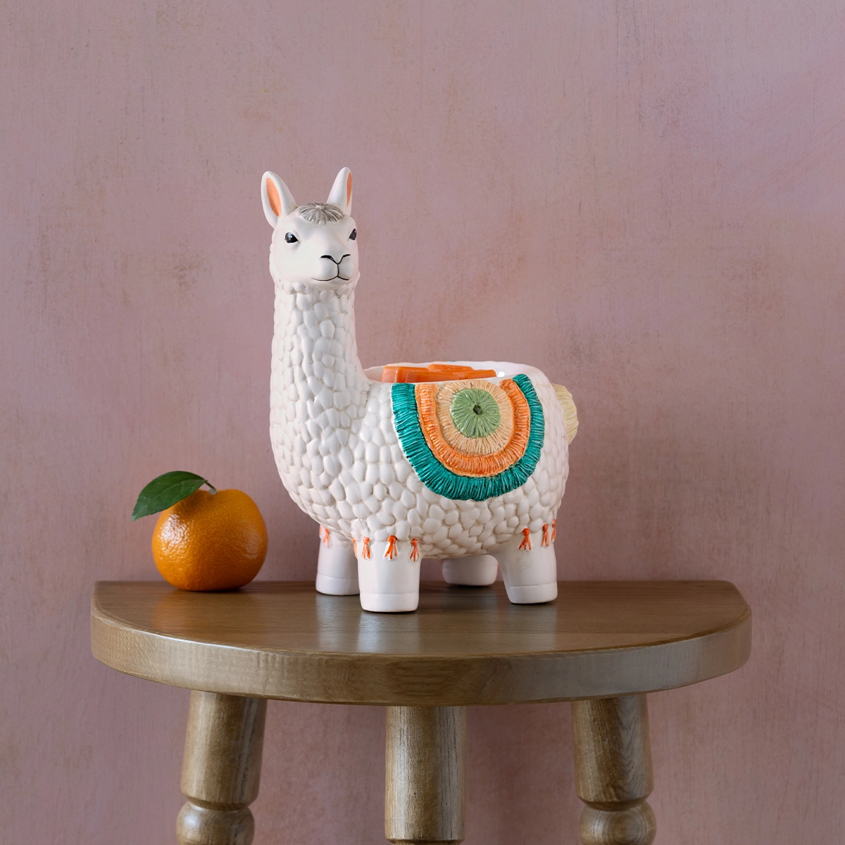 White ceramic llama warmer with textured fleece, colourful woven blanket across its back in orange, teal and green, standing on four legs with orange accents, displayed on a wooden table next to an orange