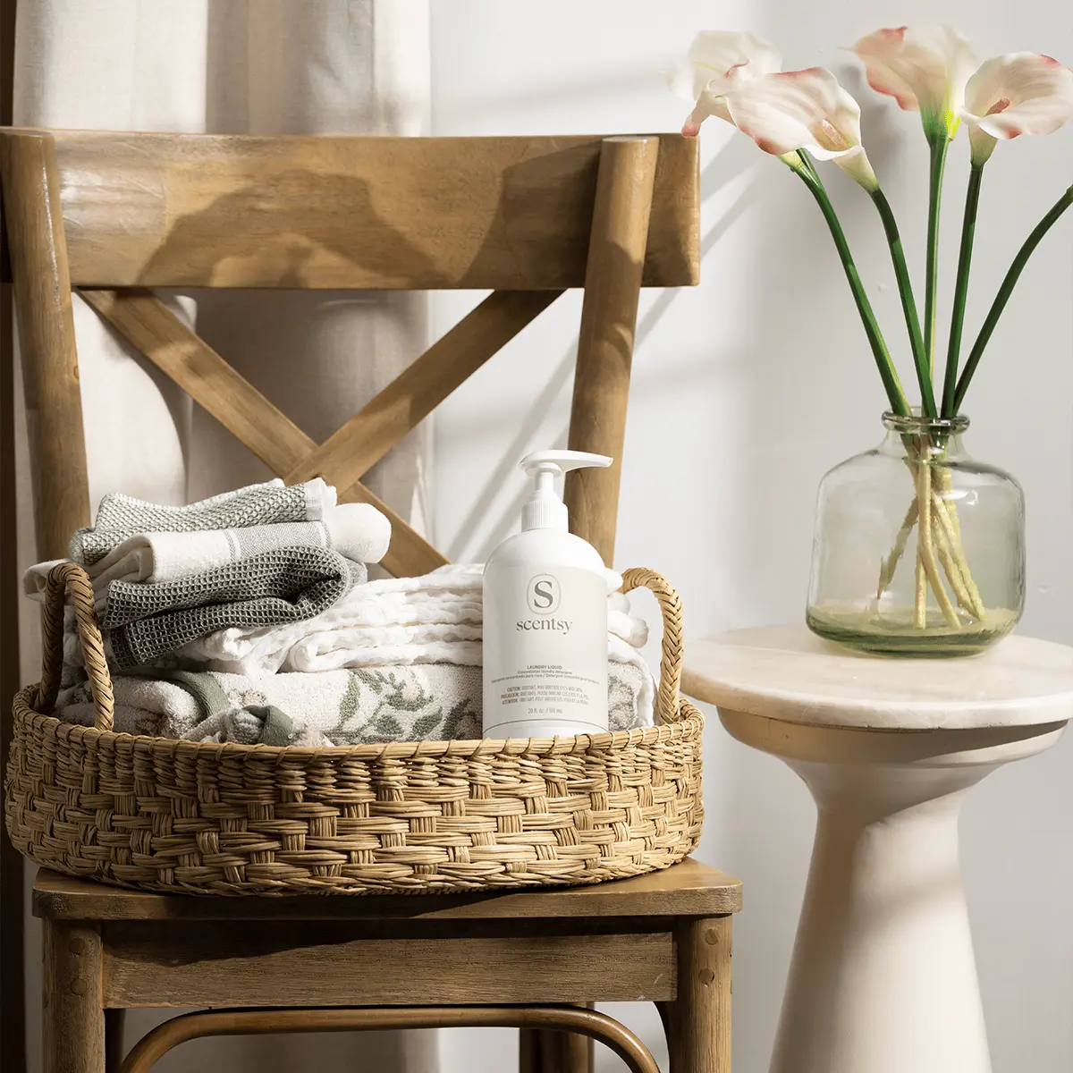 Woven basket with rope handles filled with folded grey and white towels and a white pump bottle of Magnolia Shimmer Laundry Liquid, displayed on a wooden chair