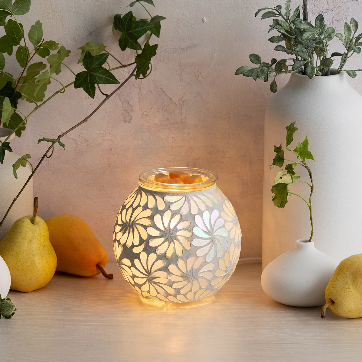 Illuminated glass Scentsy Warmer with an etched daisy pattern, the Sky Blossom design, glowing warmly on a styled surface with fresh fruit and potted plants