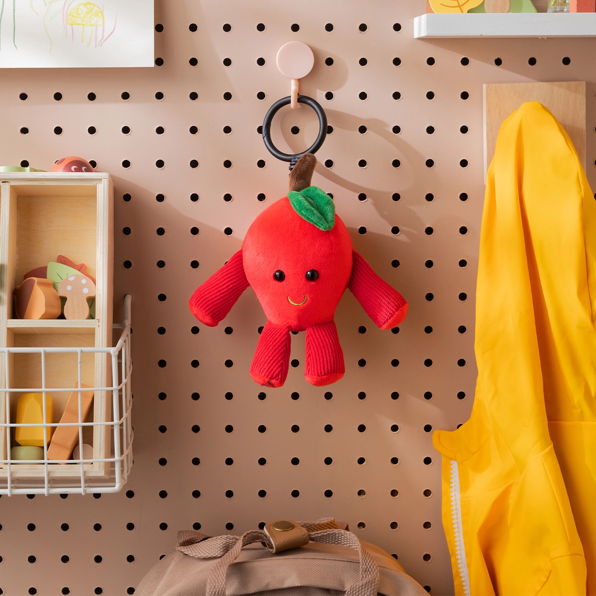 Red plush apple keychain with a smiling face, green leaf detail, and red ribbed arms and legs, hanging from a pink clip on a pegboard display