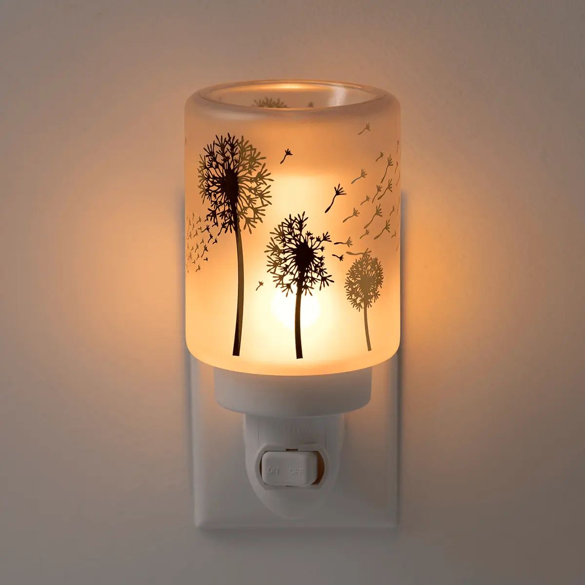 Cylindrical translucent warmer with silhouettes of dandelions and floating seeds, emitting warm amber light when switched on, mounted on a white wall plug base