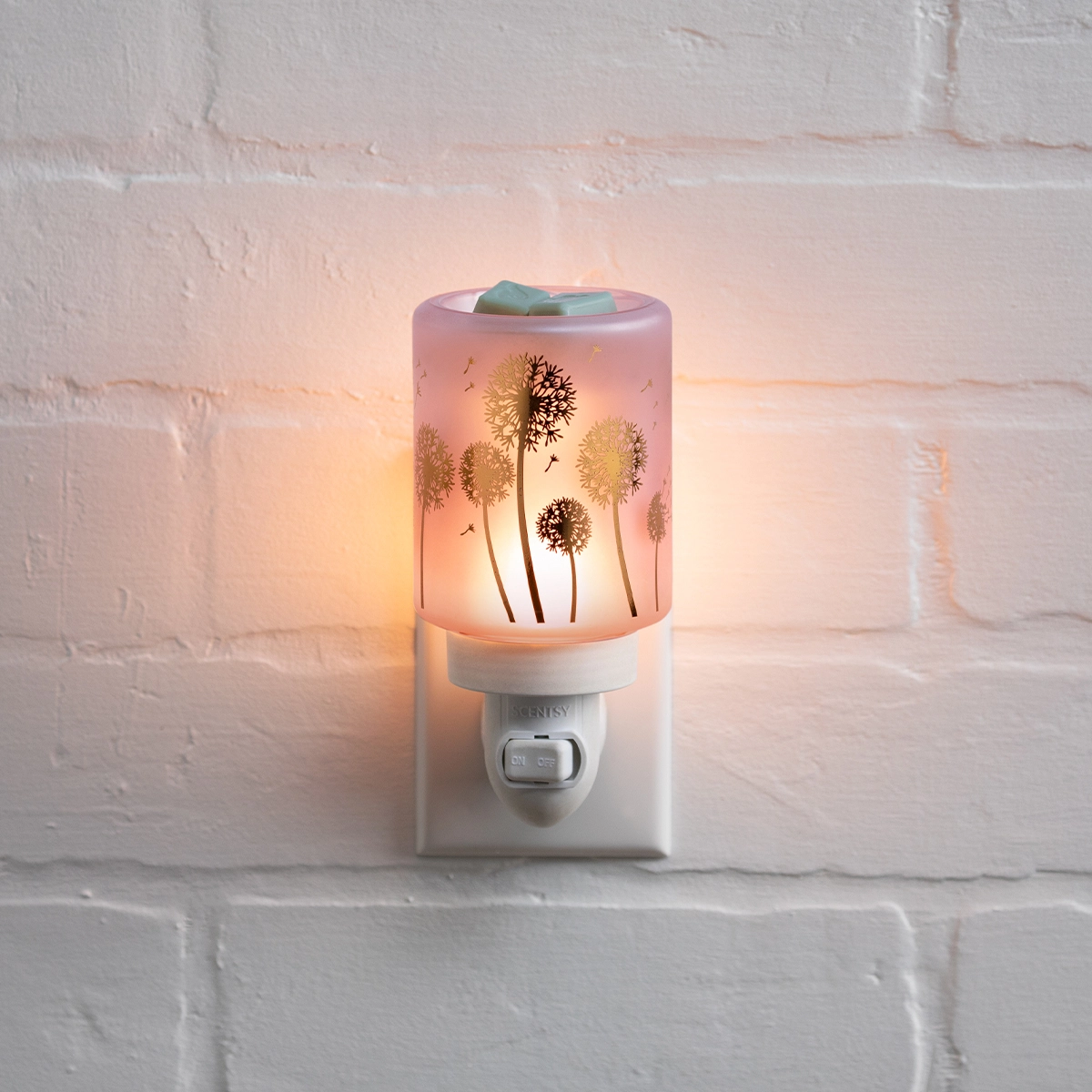 Mini Scentsy Warmer plugged into a wall outlet, featuring a cylindrical frosted glass shade with silhouetted dandelion designs and a warm glow, topped with a blue and green wax melt, the Wishbloom Mini Scentsy Warmer