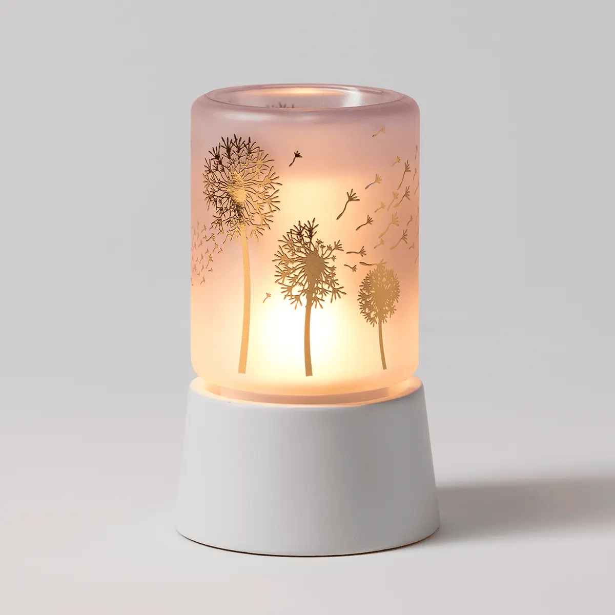 Translucent pink cylindrical warmer with etched dandelion and seedhead design, lit from within with a warm glow, seated on a white ceramic tabletop base, the Wishbloom Mini Scentsy Warmer with Tabletop Base