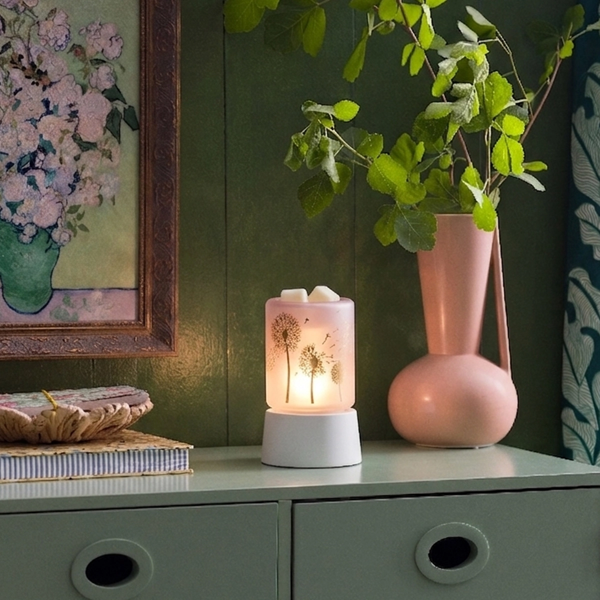 Lit mini warmer with a white base and cream ceramic top decorated with dandelion illustrations, displayed on a shelf beside a coral vase with trailing greenery