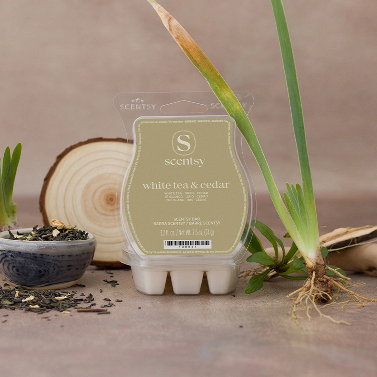Cream-coloured wax bar with a sage-green label reading "white tea & cedar", displayed in clear plastic clamshell packaging surrounded by loose tea leaves, wooden slices, and fresh green plant stems