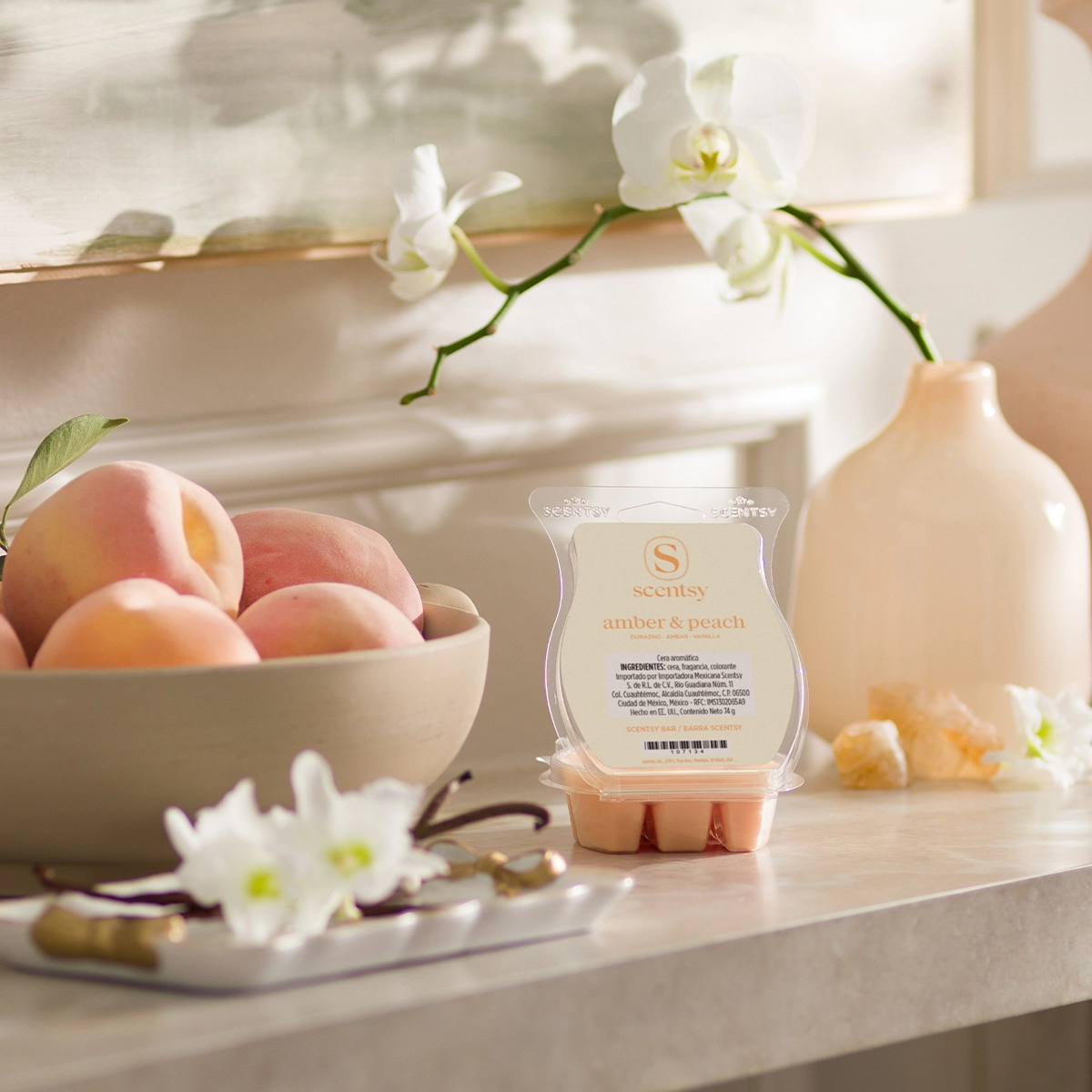 Scentsy Bar labelled Amber & Peach displayed on a bathroom counter surrounded by white orchids, peaches in a ceramic bowl, and a cream diffuser