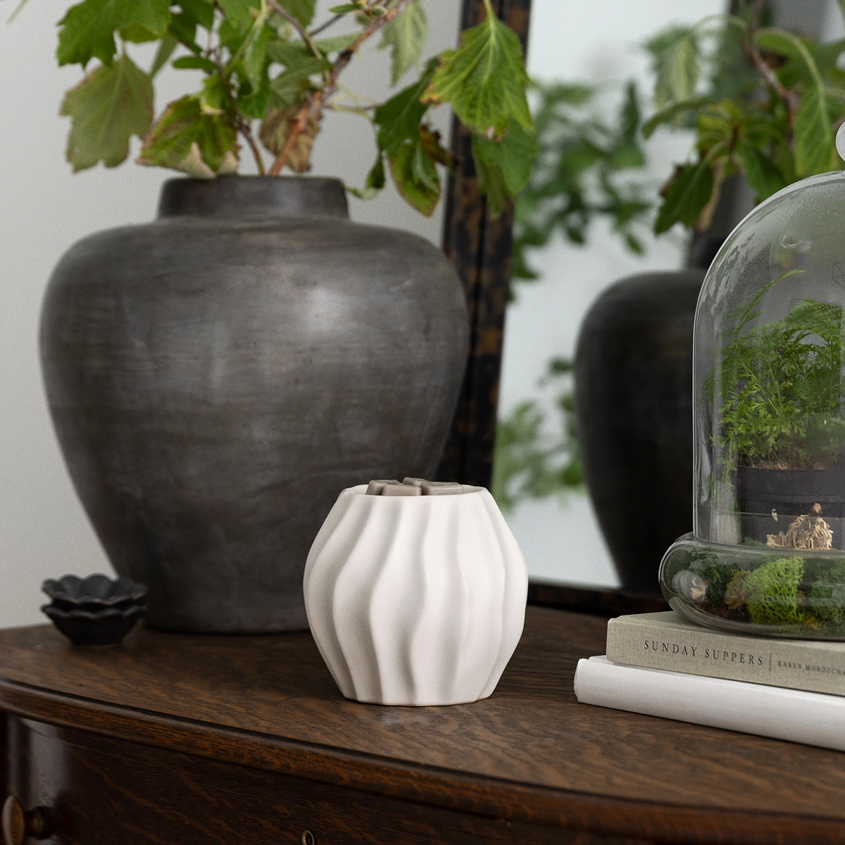 Compact white ceramic warmer with vertical ribbed detailing, displayed on a dark wooden shelf alongside black ceramic vessels and potted greenery