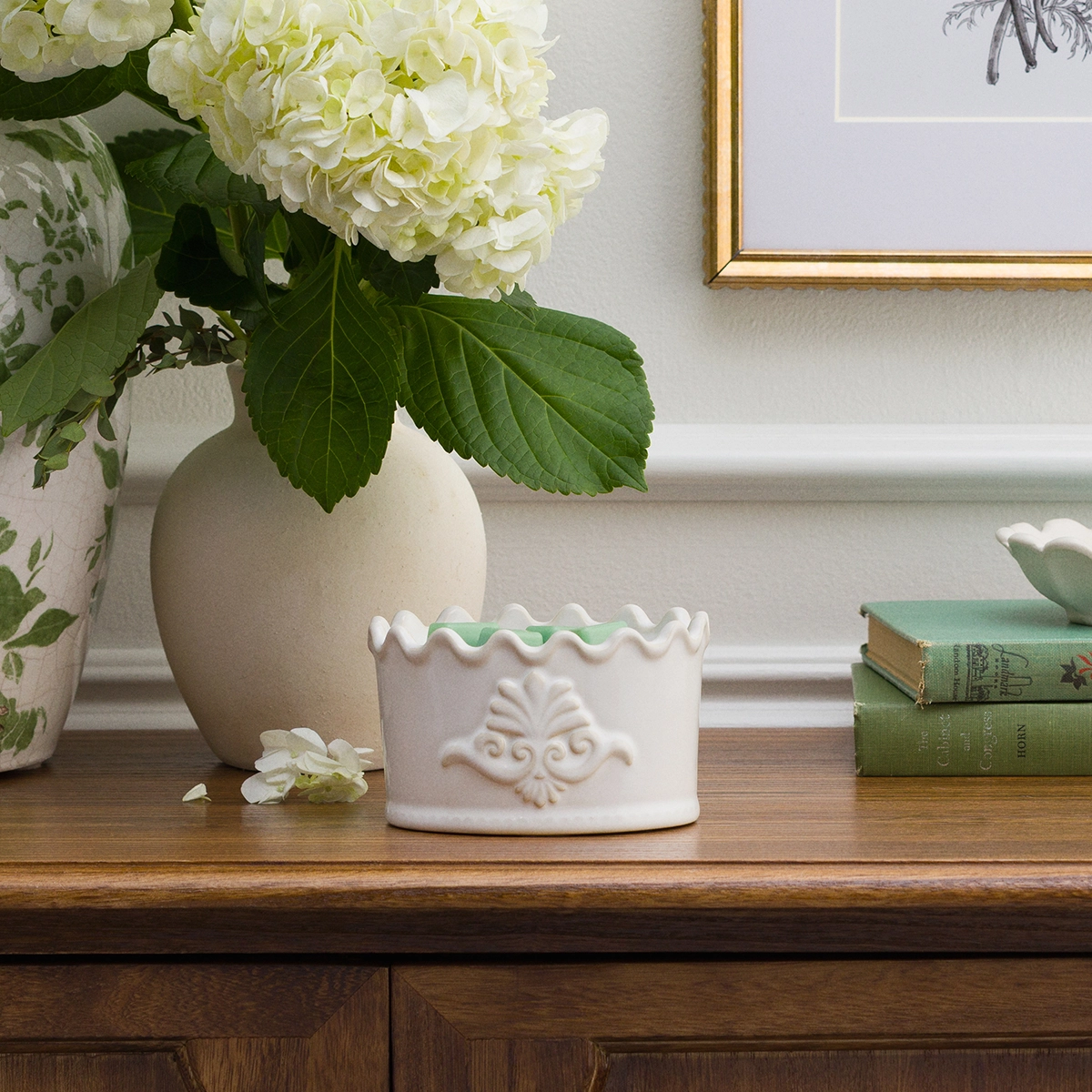 White ceramic Scentsy warmer with a scalloped rim and embossed crest motif, displayed on a wooden shelf alongside cream hydrangeas and vintage books
