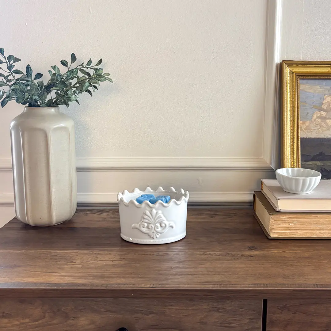 White ceramic warmer with ornate embossed crest and scalloped rim, the Classic Crest Scentsy Warmer, displayed on a wooden shelf with blue wax inside