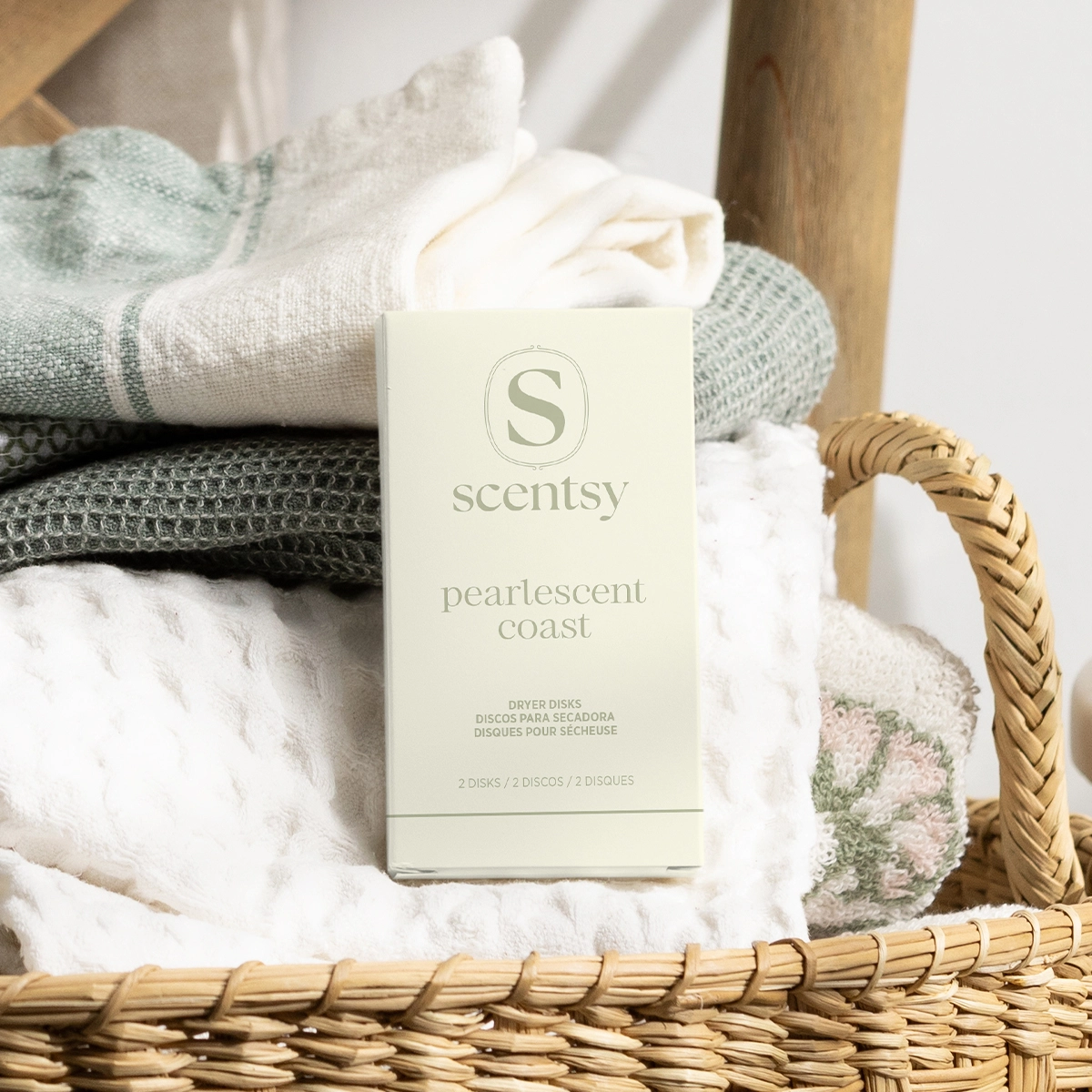 Cream-coloured box of Scentsy Pearlescent Coast Dryer Disks displayed in a woven basket with folded towels in neutral tones and a wooden chair in the background