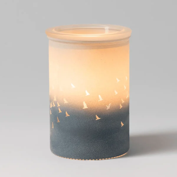 Waves & Wings Scentsy Warmer - Cylindrical wax warmer with a two-tone design: pale cream upper section and charcoal lower section with cut-out sailboat silhouettes, the Waves & Wings Scentsy Warmer, shown glowing warmly from within