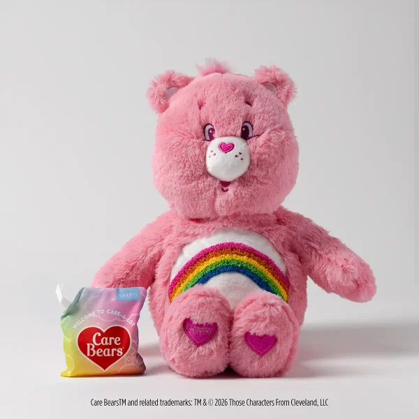 Pink plush bear with round ears, white muzzle with a heart-shaped nose, purple eyes, and a rainbow emblem on its chest with purple heart pads on its feet, the Cheer Bear Scentsy Buddy, shown with a Care Bears wax sachet