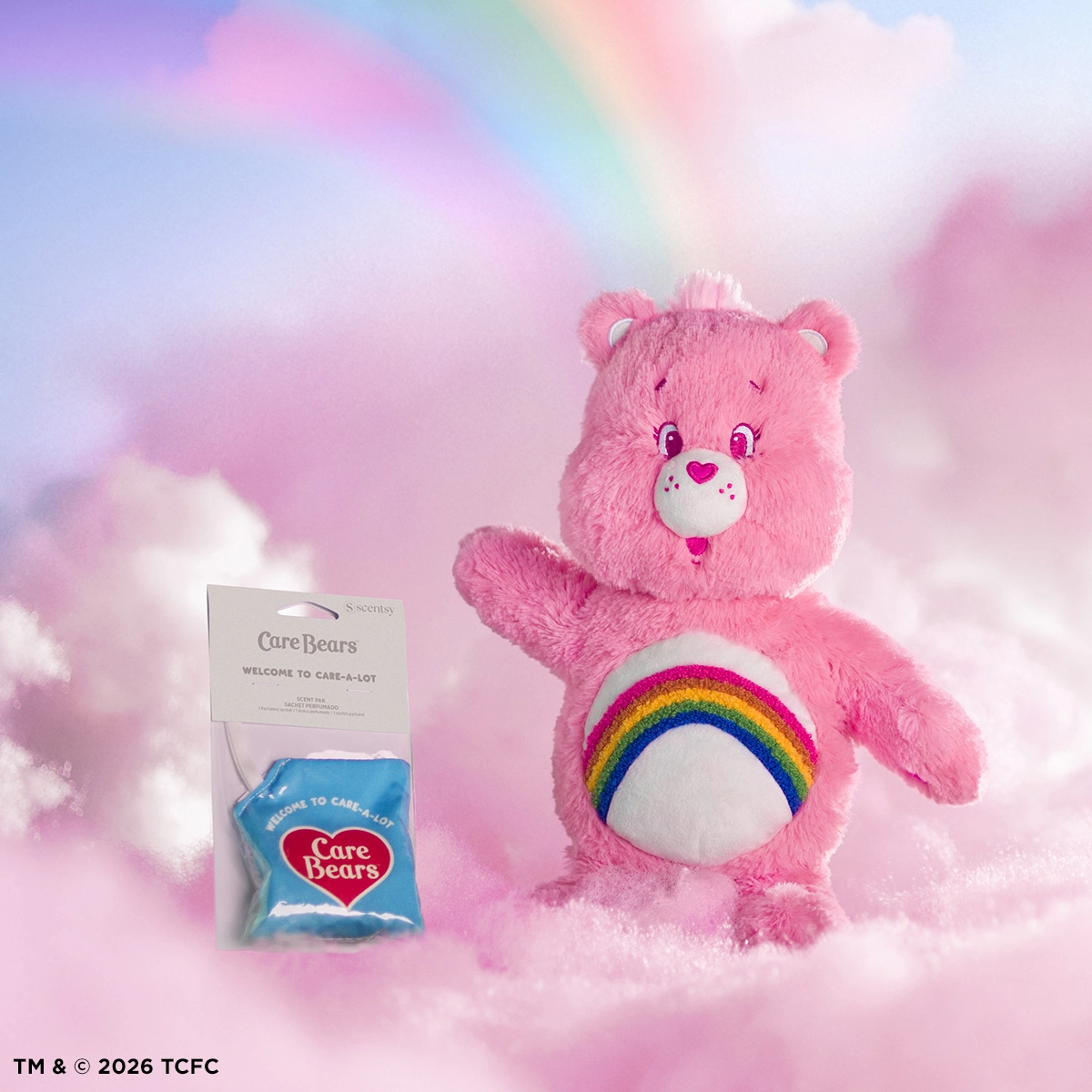Pink plush bear with a rainbow design on its belly, pink nose, and embroidered facial features, posed against a pastel cloud background alongside a blue Care Bears Scentsy Pak packet