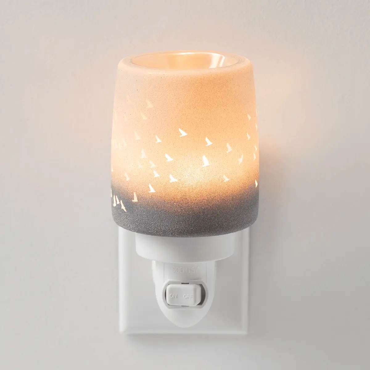 Waves & Wings Mini Scentsy Warmer - Cream and grey ceramic mini warmer with white cutout bird and wave details, lit from within, the Waves & Wings Mini Scentsy Warmer, on a white wall plug base