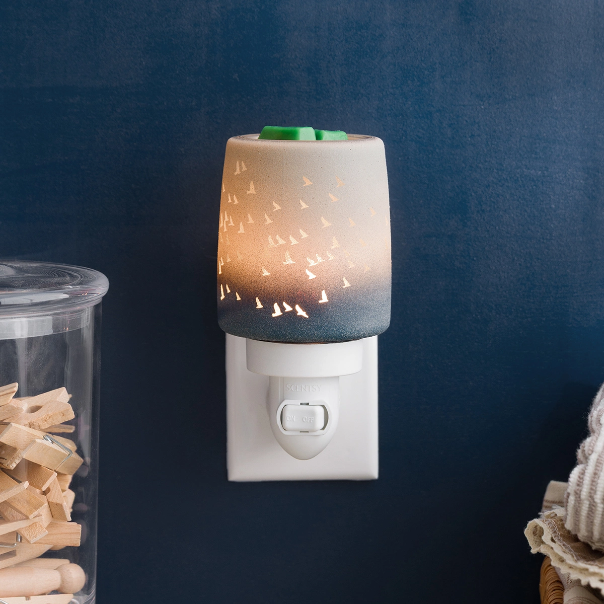 White plug-in warmer with a ceramic shade featuring a gradient from cream to deep teal, decorated with cut-out tree and bird silhouettes that cast warm light when lit, topped with green wax