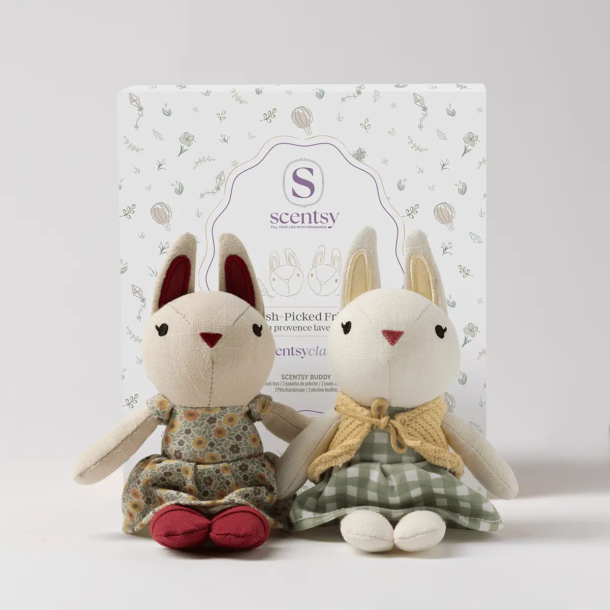 Two soft plush cat buddies with cream bodies, burgundy inner ears, and embroidered faces sit in front of their packaging, one wearing a floral print dress with red feet and the other in a green gingham skirt with a yellow bow, the Fresh-picked Friends Scentsy Buddy pair